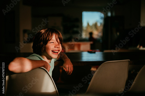 Boy sitting at a dining room table laughing with sunlight on his face