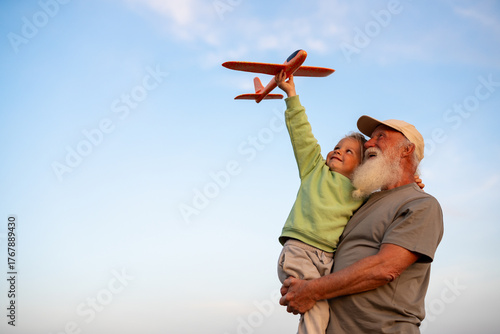 Quadro em tela Joyful grandfather holding his happy blond grandson who is lifting a toy airplane into the sky, both enjoying playful time together under a clear blue sky
