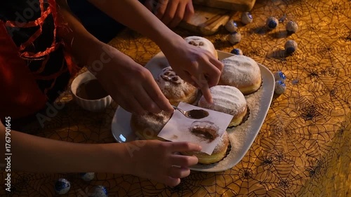 People preparing for Halloween party by decorating donuts with powdered sugar and cocoa using paper stencils, creating fun spooky faces and shapes on festive table covered with cobweb tablecloth