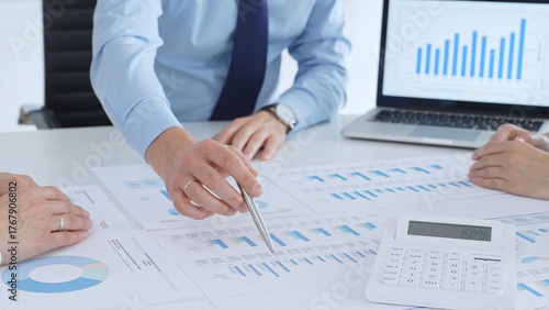 Business professional pointing at printed charts with a pen during a meeting, laptop showing bar graphs and calculator on table while team collaborates on financial analysis and planning