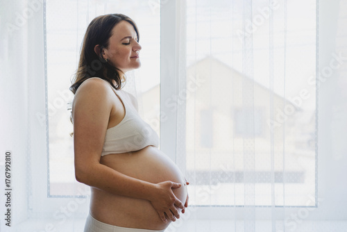 A Pregnant woman stands by a window, cradling her round belly. This moment captures the beauty of pregnancy. Free Download A Pregnant woman stands by a window, cradling her round belly. This moment captures the beauty of pregnancy. - GFXLoad