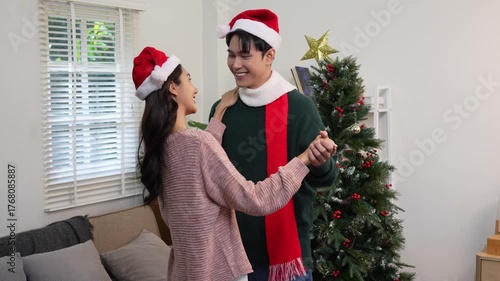 Happy Asian couple celebrating Christmas sharing warmth in winter embrace, exchanging gifts on sofa in cozy home.
