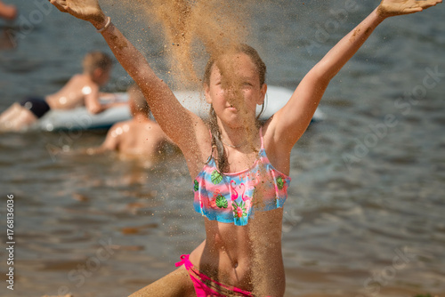 Young girl with long hair wearing colorful swimsuit stands confidently at the beach, with water glistening in the background, capturing the essence of summer fun and joy
