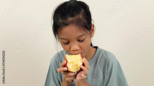 A video shows a girl standing holding a red apple and biting it bit by bit, almost halfway through. With different poses and perspectives, she smiles brightly as she enjoys eating her favourite fruit.
