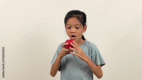 A video shows a girl standing holding a red apple and biting it bit by bit, almost halfway through. With different poses and perspectives, she smiles brightly as she enjoys eating her favourite fruit.