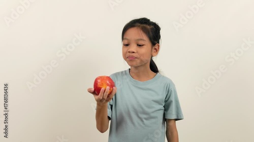 A video shows a girl standing holding a red apple and biting it bit by bit, almost halfway through. With different poses and perspectives, she smiles brightly as she enjoys eating her favourite fruit.