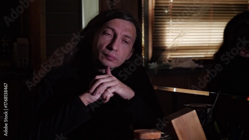 A contemplative and serious mood is captured as a man with long hair and a serious expression rests his chin on his interlocked hands in a dimly lit setting