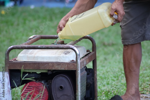 A person's muscular hands pour fuel from a plastic jerrycan into a rusty portable power generator outdoors on green grass.