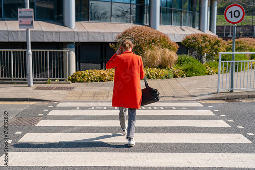 Fotografie Person in bright red coat walking across a crosswalk on a sunny day in an urban