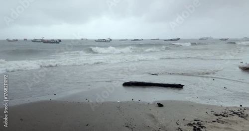 On a rainy day, choppy waves with whitecaps break on a wet, sandy shore where a dark piece of driftwood lies. In the distance, a fleet of fishing boats bobs on the rough sea under a gray, overcast sky
