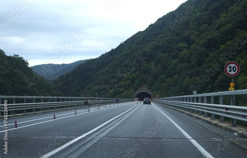 Mountain road and tunnel. A car drives along a mountain highway leading into a tunnel. The road is marked with a 30 km/h speed limit sign and barriers on both sides.