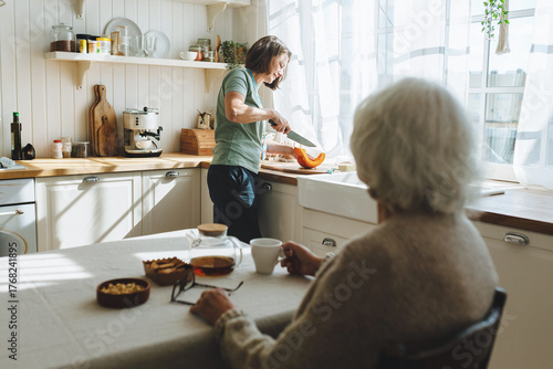 Rare view of senior lady sitting at kitchen table, having herbal tea looking at nurse or caregiver cutting pumpkin standing next to counter, having nice conversation while social worker cooking