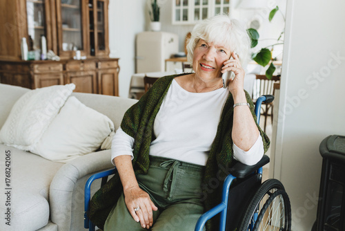 Disabled senior lady in wheelchair talking on phone with social support service, making appointment with doctor to visit her as weekly check-up at home, sitting in cozy living-room
