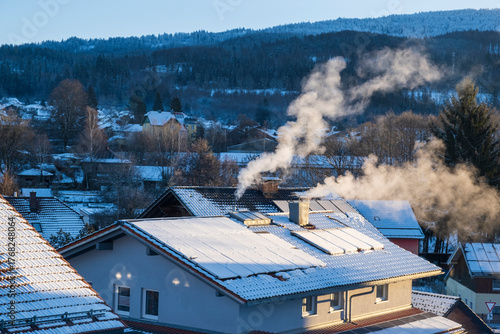 Snow-dusted rooftops, solar panels and chimneys sending smoke into cold air above a small German village on a winter morning.