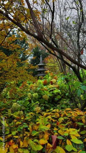 Vertical shot of a small, traditional Japanese stone pagoda or lantern partially obscured by an abundance of lush autumn foliage in varying shades of green, yellow, and faded orange, including