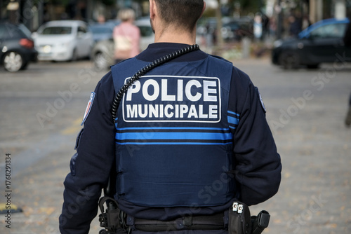 A municipal police officer seen from behind wearing a blue uniform and radio equipment while standing on a city street during daytime patrol.