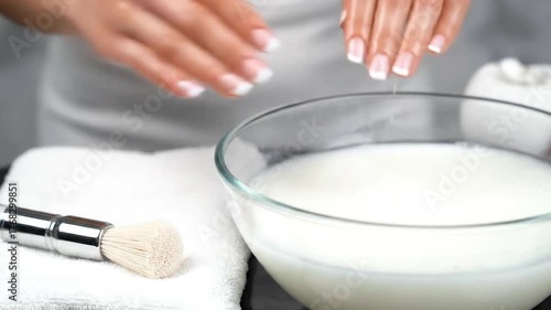 Woman Soaking Hands in Milky Water with French Manicure to Soften Skin on White Table Near White Towel and Brush in Bright Studio Lighting for Beauty and Hygiene Routines