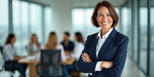 Mature businesswoman in focus with blurred team in meeting behind her in glass office, symbolizing authority, leadership, and decision-making