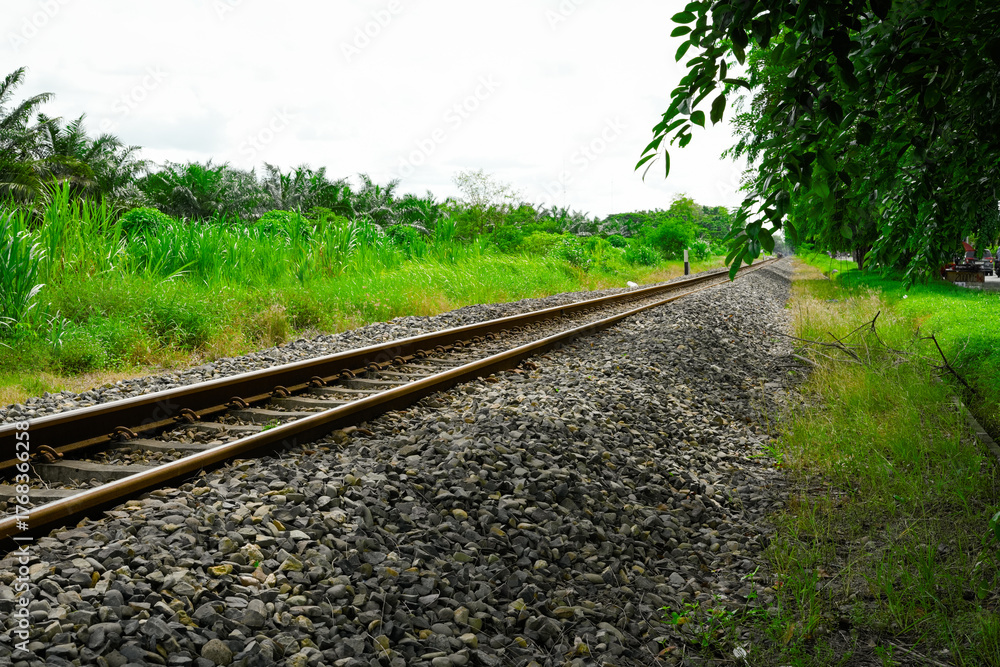 Fototapeta premium railroad tracks stretch through green rural landscape with trees, gravel bed, and open sky