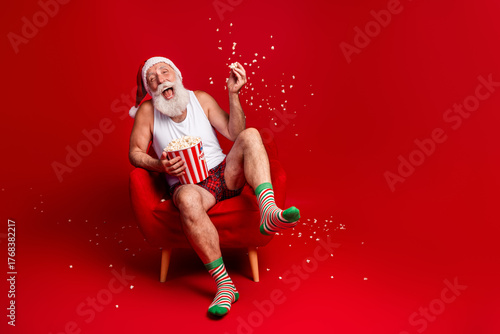 Santa enjoys popcorn on Christmas Eve relaxing in a red chair wearing hat striped socks and smiling