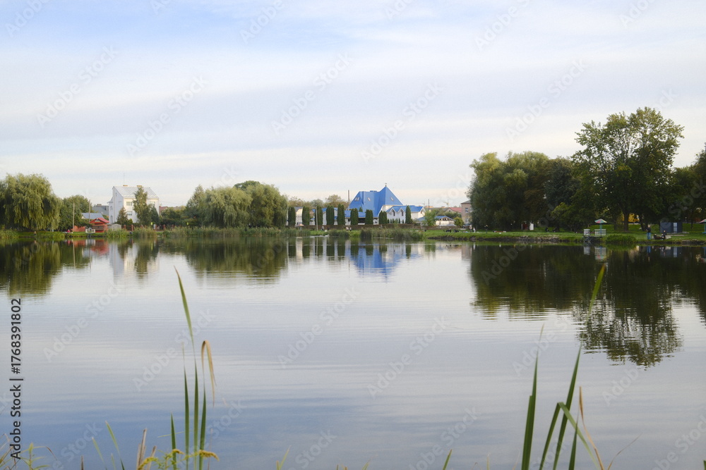 Obraz premium Calm water reflects the shoreline with buildings and trees on an overcast day.
