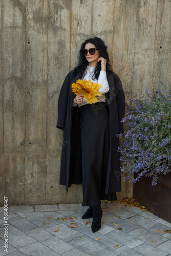 Elegant woman in sunglasses holding a bouquet of yellow autumn leaves, dressed in a white sweater and dark coat, standing against a concrete wall. Fall fashion and lifestyle concept.