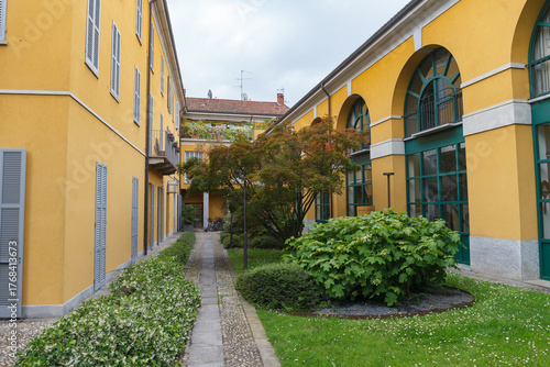 Greenery in the inner yard with pathwalks, historical stone bulding, yellow color, yearly XX century. Monza, Italy.