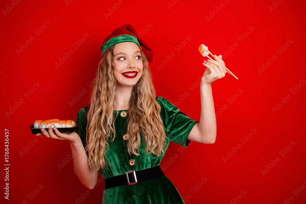 Fototapeta premium Festive young elf in green outfit holding sushi with stylish smile against vibrant red background celebrating holiday joy