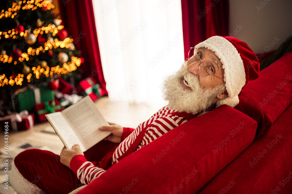 Obraz premium Elderly man in Santa outfit reading indoors by a decorated Christmas tree and enjoying the festive holiday atmosphere