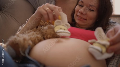 Pregnant woman and her husband embrace on the couch, sharing a loving moment while he gently touches her belly with baby shoes.