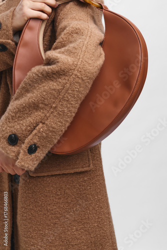 Quadro su tela Asian woman wearing a warm brown coat stands with a stylish leather bag, presenting a chic urban look that conveys confidence, elegance, and contemporary fashion in a minimal setting