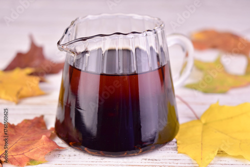 Maple syrup in a glass gravy boat against a backdrop of autumn maple leaves.
