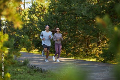 Young adult Caucasian man and young adult Caucasian woman jogging together on wooden path surrounded by green trees, both maintaining steady pace and focused expressions outdoors