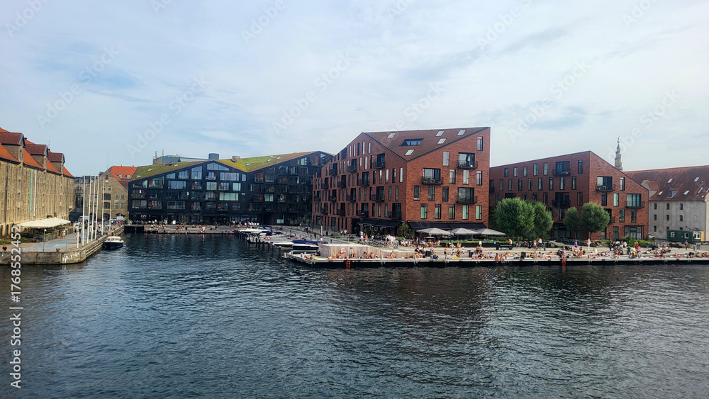 Naklejka premium 07 August, 2024, Copenhagen city center, Denmark Summer day scenic canal with boats and tourists, cyclists riding along colorful buildings and waterfront, vibrant urban atmosphere in European capital