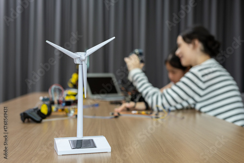 Close-up of wind turbine model with blurred girls working on robotics in background, representing renewable energy, STEM education and future technology innovation.