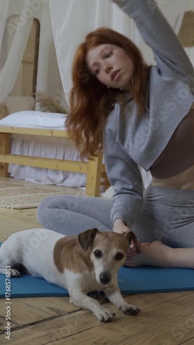 Young beautiful woman doing sport stretching exercises. Cute small dog resting next to her on blue mat. Healthy lifestyle vertical video footage. Sport with pets at home following video online class 