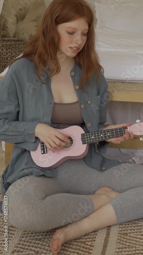beautiful red-haired girl with long hair in blue shirt sits on the floor in the living room at home, learning to play ukulele. relaxed hobby time. following video lesson online. vertical video footage