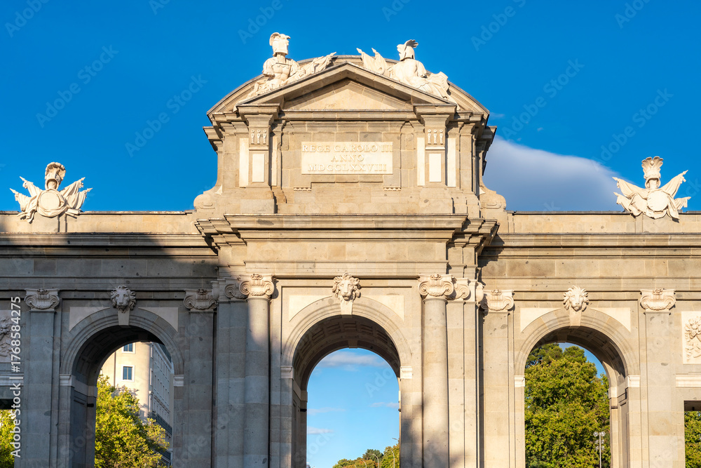 Obraz premium Puerta de Alcalá stands tall under afternoon sunlight in Madrid, Spain. The blue sky and shadows enhance its neoclassical design and sculptural details, framed by greenery at Plaza de la Independencia