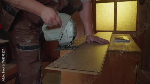 Skilled male worker in uniform using an electric jigsaw to accurately cut a wooden laminate board for floor installation during a home renovation or new construction project in a workshop.