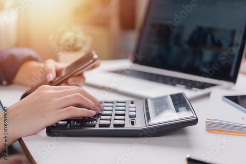 Woman hand use calculator, holding smartphone, work office desk laptop. Financial accounting concept. Calculate tax, budget, data analysis, expense report. remote work, multitasking, sunlight.
