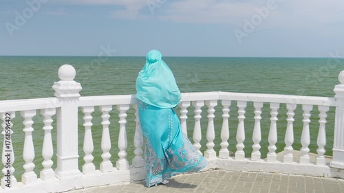 A young Muslim woman in blue stands on the embankment by the railing, looking out to sea. General shot.
