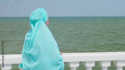 A young Muslim woman in blue stands on the embankment by the railing, looking out to sea. Medium shot.