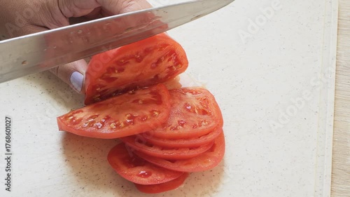 A woman's hand slices a ripe red tomato into rings on a board. Medium shot.
