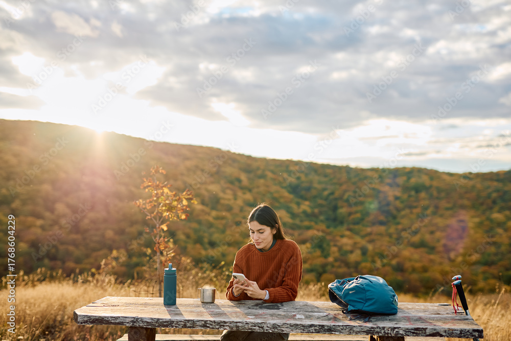 © we.bond.creations - Woman taking a break while hiking, sitting at a picnic table and using phone during sunset in the mountains © we.bond.creations - Woman taking a break while hiking, sitting at a picnic table and using phone during sunset in the mountains