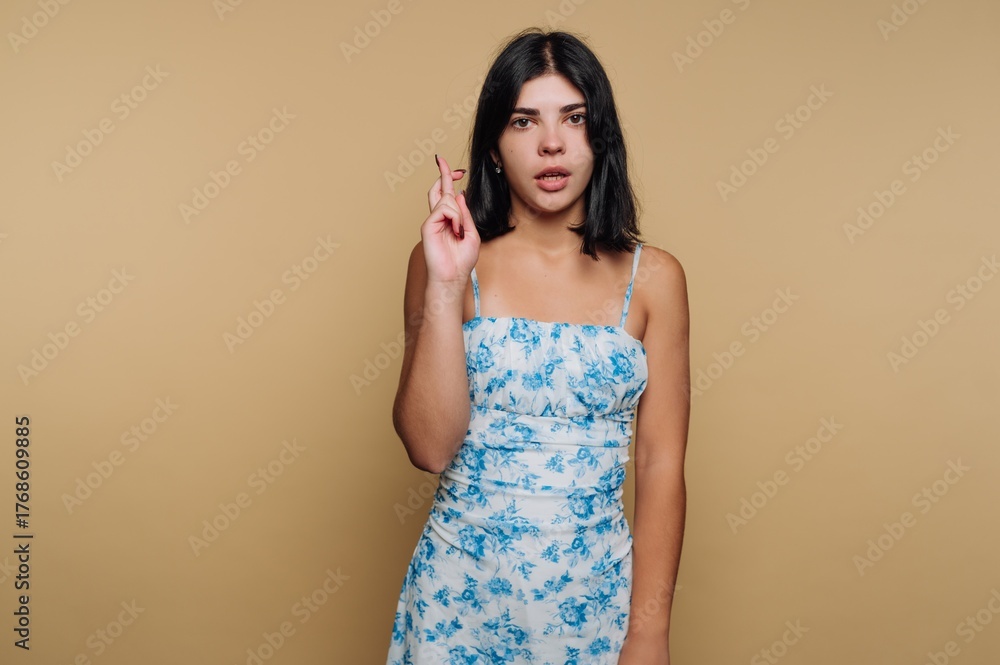 Obraz premium Young woman in floral dress crossing fingers on beige background.