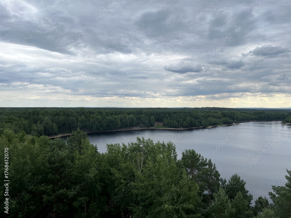 Fototapeta premium Scenic view of a calm river surrounded by green forest under a cloudy sky. Peaceful natural landscape perfect for concepts of tranquility, ecology, and outdoor adventure.