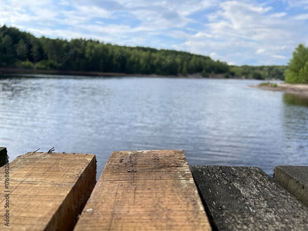 Naklejka premium Wooden pier by a calm river on a sunny day. Tranquil nature landscape with water, forest, and blue sky. Perfect for concepts of relaxation, travel, and summer outdoors.