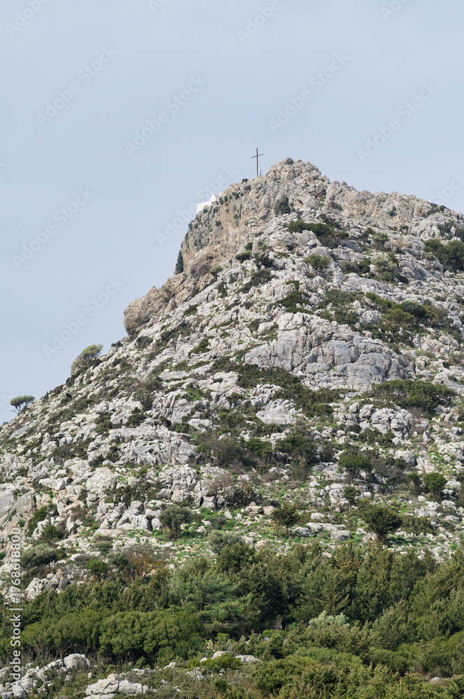 Fototapeta premium Rocky mountain peak with cross and small chapel surrounded by greenery in Crete