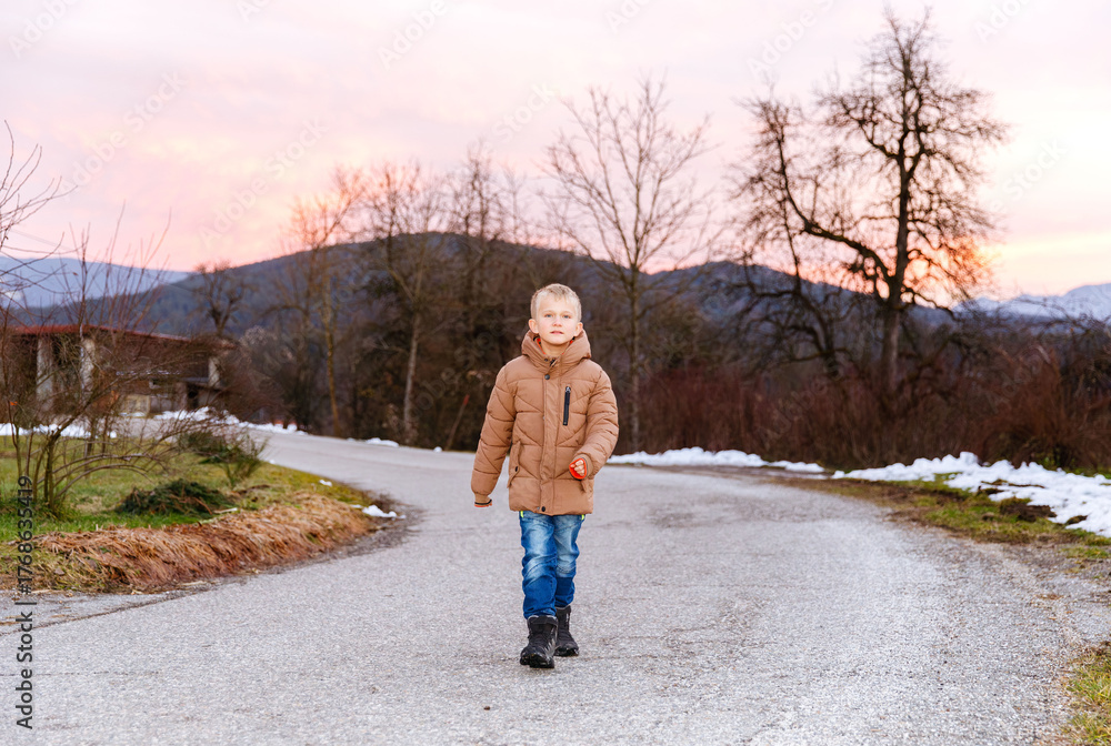 Fototapeta premium Caucasian boy walking on country road at sunset in winter landscape