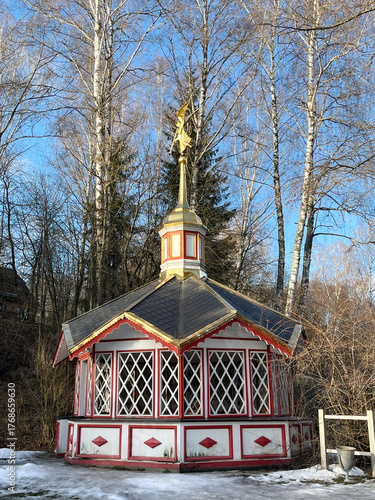 Colorful octagonal gazebo with ornate details and a golden spire, surrounded by tall trees and snow, showcasing architectural beauty in a serene outdoor setting